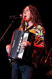 Una fotografía de "Weird Al" Yankovic, cantando a través de un micrófono y tocando una armónica.
