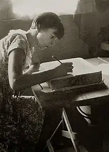 full length portrait of young woman with short hair seated working with a pencil on a litho stone