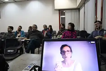 En el primer plano de la imagen se ve una laptop sobre la mesa con la imagen de Margarita Padilla en primer plano. Por detrás y un poco difuso un grupo de personas siguen atentas su presentación..