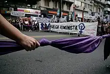 Mujeres reunidas en una gran ronda en la calla. Al frente, sostienen un pañuelo violeta anudado. Al fondo una pancarta que dice huelga feminista.