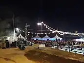 Se aprecia desde arriba en el lado izquierdo cajas del grupo de música, al fondo unas carpas blancas y una gran cantidad de gente bailando. Destacan de la imagen las luces que envuelven la verbena